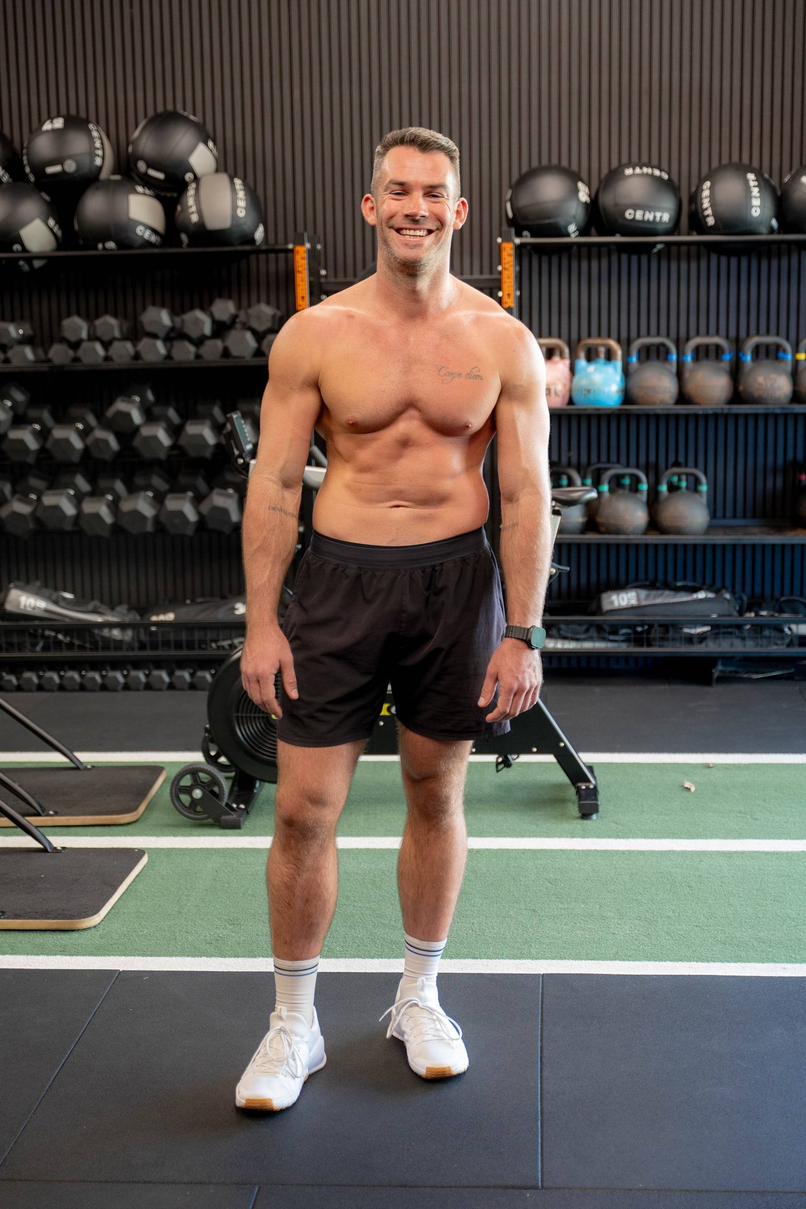 Richy standing in a gym in front of a rack of kettlebells, smiling